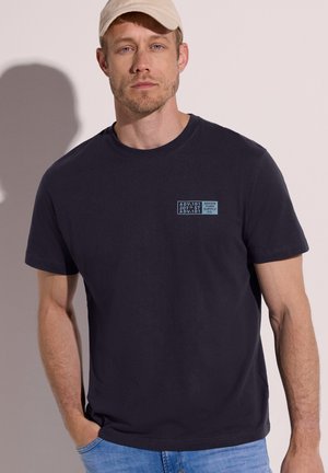 Mand iført beige kasket, sort t-shirt med lyseblå tekst på brystet og blå jeans står mod en ensartet lys baggrund.