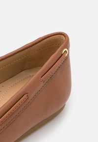 Chaussure en cuir marron à enfiler avec une finition lisse, accent en métal doré et bout rond. L'intérieur est dotée d'une rembourrage beige.