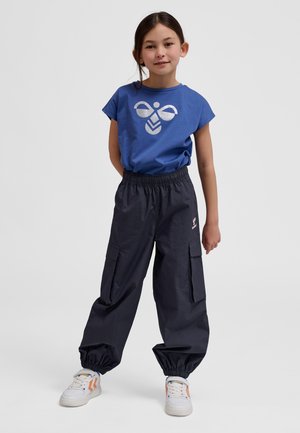 Κορίτσι που στέκεται φορώντας μπλε μπλουζάκι με ασημένιο λογότυπο μέλισσας, μαύρο παντελόνι cargo με ελαστικά μανσέτες και λευκά αθλητικά παπούτσια με πορτοκαλί λεπτομέρειες.