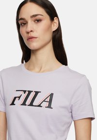 Camiseta de algodón lila con un atrevido gráfico del logotipo "FILA" en negro y rojo en la parte frontal, con cuello redondo y mangas cortas.