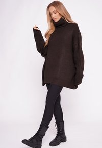 Übergroßer, dunkelbrauner Pullover mit hohem Rollkragen und geripptem Material, kombiniert mit schwarzen Leggings und Schnürkampfstiefeln.