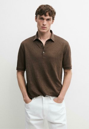 Junger Mann mit kurzen lockigen braunen Haaren, der ein braunes kurzärmeliges Poloshirt und weiße Hosen trägt und die Hände in den Taschen hat.