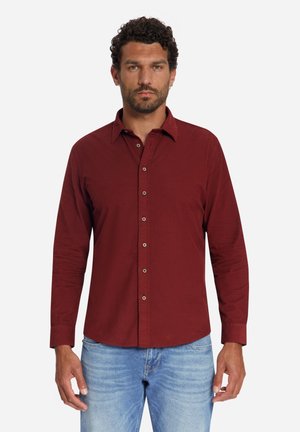 Mann mit lockigem Haar, der ein bordeauxfarbenes, langärmliges Hemd mit Knöpfen und hellblaue Jeans trägt, steht vor einem einfarbigen Hintergrund.