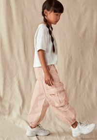 Witte, kortemouw t-shirt gecombineerd met lichtroze cargobroek met zijzakken en elastische boorden, aangevuld met witte sportieve sneakers.