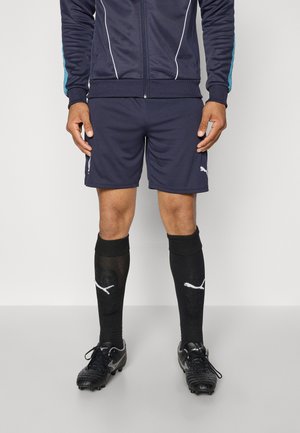 Marine sportsshorts med hvite detaljer, parret med svarte kompresjonsstrømper med logo, og svarte atletiske fotballsko.