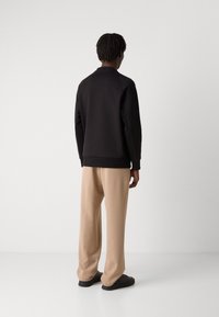 Μαύρη μπλούζα με ψηλό λαιμό και ριγωτό τελείωμα, συνδυασμένη με χαλαρές μπεζ παντελόνες και μαύρα αθλητικά παπούτσια, από την πίσω πλευρά.