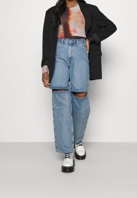 Breda jeans med rivningar i ljusblå denim, parat med en grafisk långärmad topp och en svart kavaj. Skorna består av vita plattform stövlar.