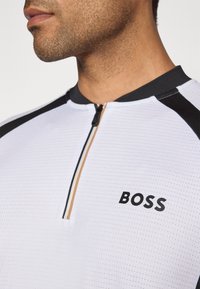 Nahaufnahme eines Mannes in einem weißen Sporthemd mit Reißverschluss, schwarzem Kragen, schwarzen Schulterstreifen und dem Text "BOSS" auf der Brust.
