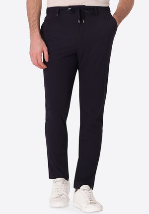 Uomo che indossa pantaloni neri aderenti con vita con coulisse e sneakers bianche, in piedi con una mano in tasca.