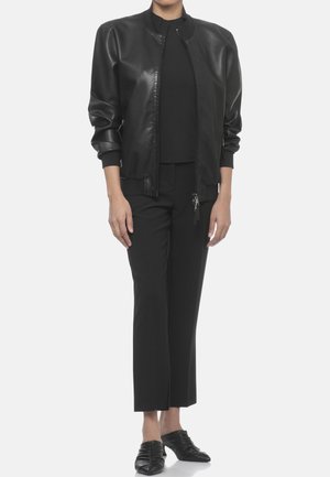 Persona che indossa una giacca bomber in pelle nera sopra un top nero, pantaloni neri corti e mule nere con punta stretta e tacco basso.