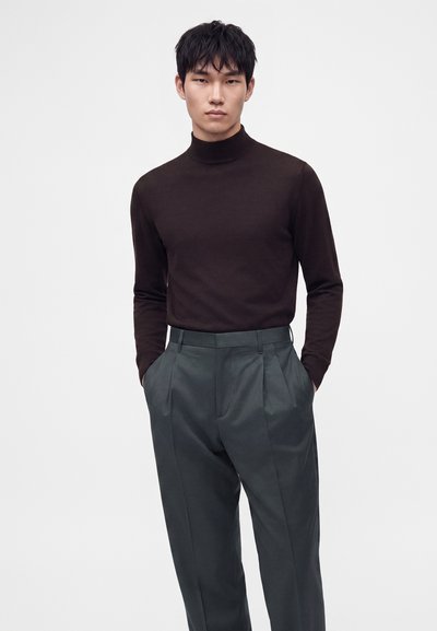 Jeune homme portant un col roulé marron foncé ajusté et un pantalon plissé gris taille haute, les mains dans les poches devant un fond uni.