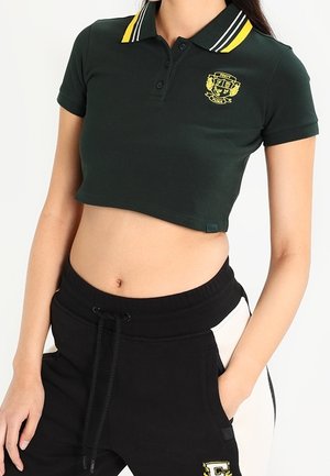Woman wearing a dark green cropped polo with yellow-striped collar and black sweatpants with white side panels and drawstring waist.