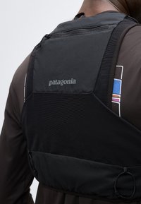 Άτομο που φοράει μαύρο τσαντάκι Patagonia στο στήθος με τσέπη με φερμουάρ πάνω από σκουρόχρωμο μακρυμάνικο πουκάμισο με πολύχρωμο γραφικό στο μανίκι.