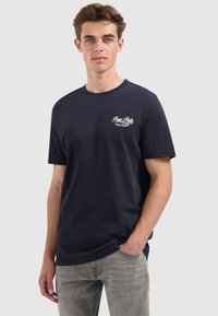 Jonge man met kort bruin haar, gekleed in een donkerblauwe "Pure Path" t-shirt en lichtgrijze jeans, staande met één hand in zijn zak.