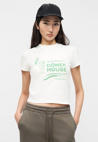 Νεαρή γυναίκα που φορά λευκό κομμένο T-shirt με πράσινο γραφικό «Döner House», μαύρο καπέλο και γκρι φόρμα με τα χέρια στις τσέπες.