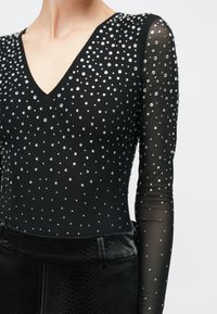 Μαύρο κορμάκι με ντεκολτέ σε σχήμα V, μακριά διάφανα μανίκια, διακοσμημένο με σκορπισμένα στρας. Υφασμάτινες μαύρες λεπτομέρειες με υφή φιδιού στη μέση.