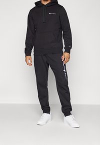 Sweat-shirt noir à capuche avec poche avant, arborant un petit logo. Associ é à un pantalon de survêtement noir affichant le texte de la marque en blanc. Baskets blanches.