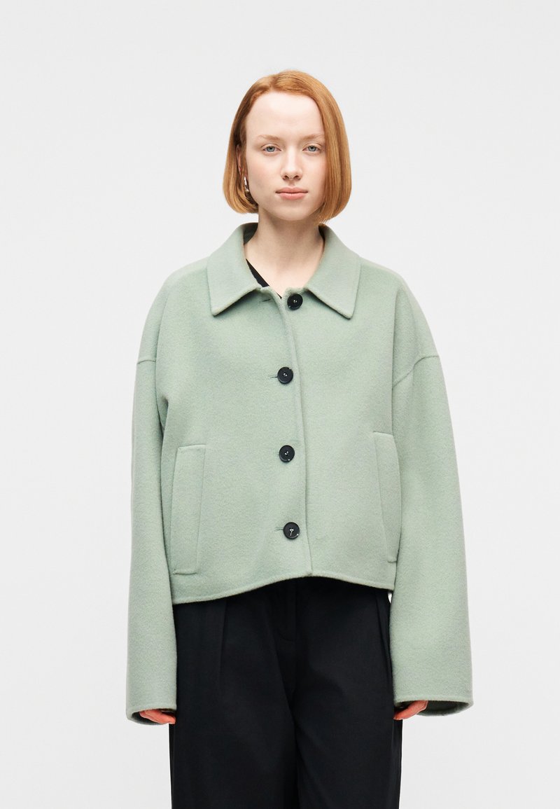 Νεαρός με κοντά κόκκινα μαλλιά, ντυμένος με ανοιχτό πράσινο πανωφόρι με κουμπιά και μαύρο παντελόνι, μπροστά σε απλό ανοιχτό φόντο.