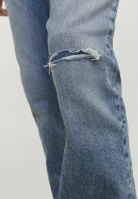 Jeans de denim azul claro con una rodilla desgastada, que presentan bordes deshilachados y una textura ligeramente desvanecida. Diseño de pierna recta con un aspecto casual.