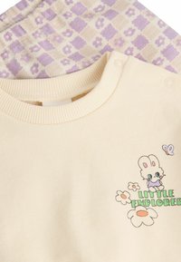 Hanorac de culoare crem, cu guler ribat, având un grafic cu un iepure și textul "Mic Explorator". Fundalul este decorat cu un model în nuanțe de lila și crem.