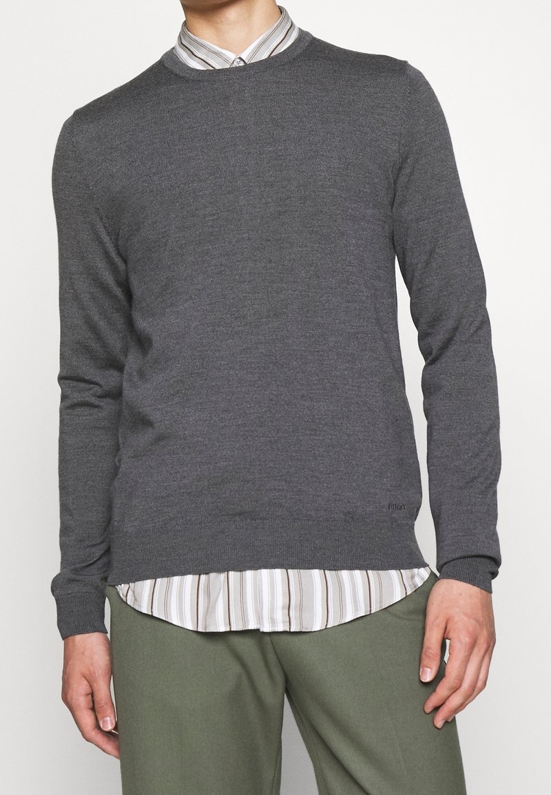 Grijze gebreide trui met lange mouwen en een ronde hals, gedragen over een wit en grijs gestreepte blouse met knoopsluiting, gecombineerd met groene broek.