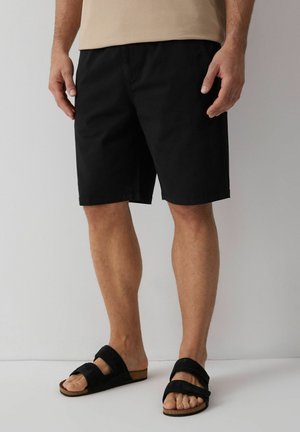 Mężczyzna noszący czarne krótkie spodenki do kolan oraz czarne sandały na dwóch paskach, stojący na tle jednolitego jasnego tła.