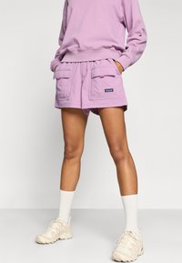 Ανοιχτό μωβ σορτς από απαλό ύφασμα με δύο εμπρόσθιες τσέπες και ελαστική μέση. Συνδυάζεται με φούτερ και μπεζ αθλητικά παπούτσια.