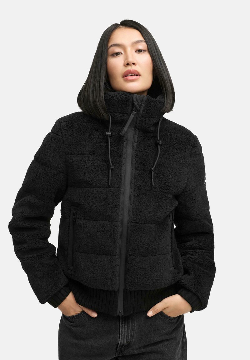 Mujer con pelo negro liso vistiendo una chaqueta de forro polar negra con cremallera y cuello alto, con las manos en los bolsillos, frente a un fondo blanco.