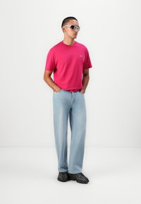 Lyserød kortærmet t-shirt med et lille logo, lysblå wide-leg jeans og sorte sneakers. Modellen bærer grå solbriller.