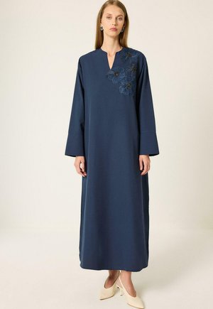 Femme portant une longue robe bleu marine à larges manches avec une broderie florale sur la poitrine, associée à des escarpins crème.