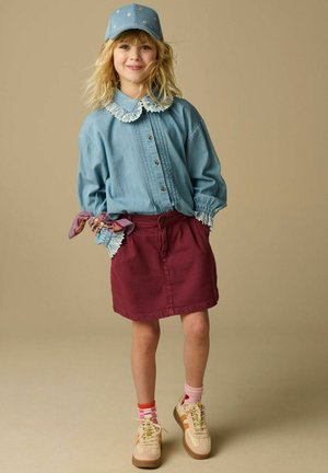 Jeune fille souriante portant un chemisier bleu à boutons avec col en dentelle, une jupe bordeaux, une casquette bleue, des chaussettes dépareillées et des baskets beiges.