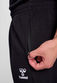 Pantalones deportivos negros con un bolsillo con cremallera, cinturilla elástica y detalle de logo en blanco. Tejido suave con un ajuste holgado y costuras curvas.