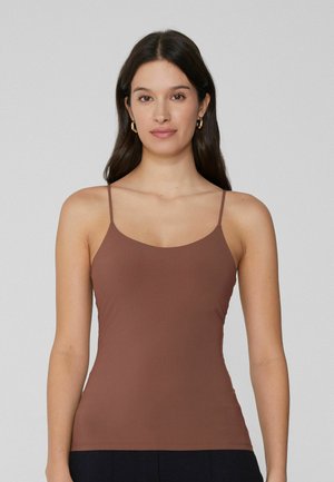 Frau mit langen, dunklen Haaren, die goldene Creolen und ein eng anliegendes braunes Camisole-Oberteil trägt, vor einem schlichten hellgrauen Hintergrund stehend.