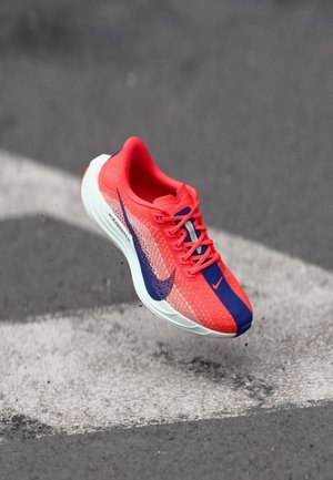 Chaussure de course rouge vif et bleue avec une semelle blanche, en l’air au-dessus de l’asphalte avec un marquage au sol blanc.