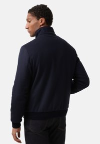 Ein Mann mit lockigem Haar, der eine dunkelblaue Jacke und dunkle Jeans trägt, von hinten gegen einen schlichten hellen Hintergrund gezeigt.
