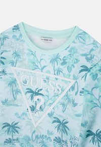 Maglietta a maniche corte azzurro chiaro con motivo tropicale a palme, caratterizzata da un logo triangolare bianco con il testo "GUESS?" al centro.