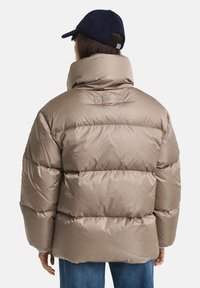 Pufferjack in lichtbruin, met een hoge kraag, een geplooid design en een gladde textuur. Model draagt een donkere pet en een blauwe spijkerbroek.