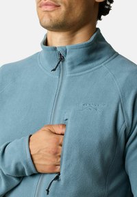 Giacca in pile di colore azzurro chiaro con chiusura a zip, colletto alto e logo ricamato. Presenta una tasca sul petto con zip e dettagli elastici ai polsi.