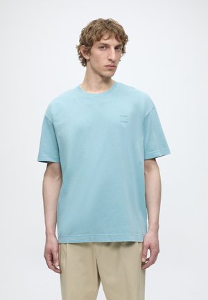 Młody mężczyzna z krótkimi kręconymi włosami, ubrany w jasnoniebieski oversize T-shirt i beżowe spodnie na jednolitym białym tle.