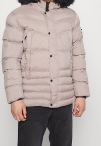 Jasno beżowa puchowa kurtka z odpinanym futrzanym kapturem, pikowany wzór i zapięcia na napy z przodu. Posiada boczne kieszenie na zamek.