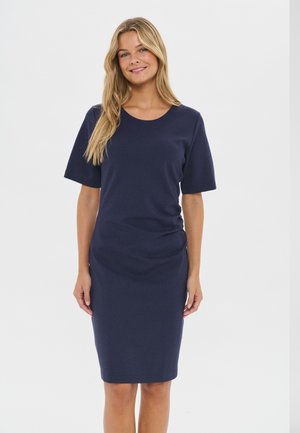 Femme aux longs cheveux blonds portant une robe bleu marine mi-longue à manches courtes, se tenant devant un arrière-plan clair uni.
