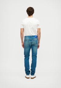 Στενά μπλε τζιν παντελόνια με ελαφρώς ξεθωριασμένο σχέδιο, πίσω τσέπες και μπλε ετικέτα HUGO στη ζώνη. Συνδυασμένα με ένα απλό άσπρο t-shirt και άσπρα αθλητικά παπούτσια.