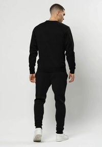 Man die een zwart sweatshirt met lange mouwen en zwarte joggerbroek draagt, staand en met de rug naar de kijker op een effen witte achtergrond, met witte sneakers.