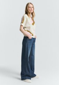 Haut en tricot avec une base crème clair, présentant des motifs abstraits colorés en rose et marron, associé à un jean en denim bleu à jambes larges.