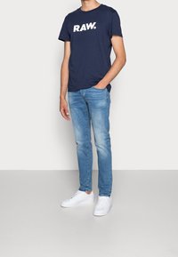 Mörkblå bomulls t-shirt med "RAW." logga i vitt, kombinerad med ljusblå jeans och vita sneakers. Avslappnad outfit med en bekväm passform.