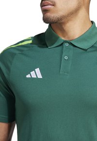 Polo verde realizzato in tessuto testurizzato. Presenta una chiusura con due bottoni, maniche corte e un logo Adidas bianco sul petto.