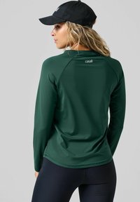 Ciemnozielona koszula z długim rękawem o gładkiej fakturze, z okrągłym dekoltem i raglanowymi rękawami. Logo na plecach w pobliżu dekoltu.