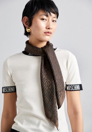 Personne aux cheveux courts et noirs porte un haut blanc Moschino, une écharpe Moschino marron nouée autour du cou et de grosses boucles d'oreilles dorées en forme d’ourson.