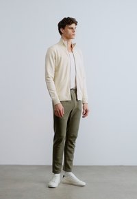 Vaaleanbeige vetoketjullinen takki, valkoinen istuva paita, oliivinvihreät housut ja valkoiset lenkkarit. Minimalistinen muotoilu, sileät tekstuurit ja rento istuvuus.