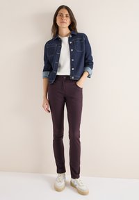 Džínová bunda tmavě modré barvy se dvěma předními kapsami, bílá strukturovaná košile, burgundské slim-fit kalhoty a bílé tenisky s modrými akcenty.